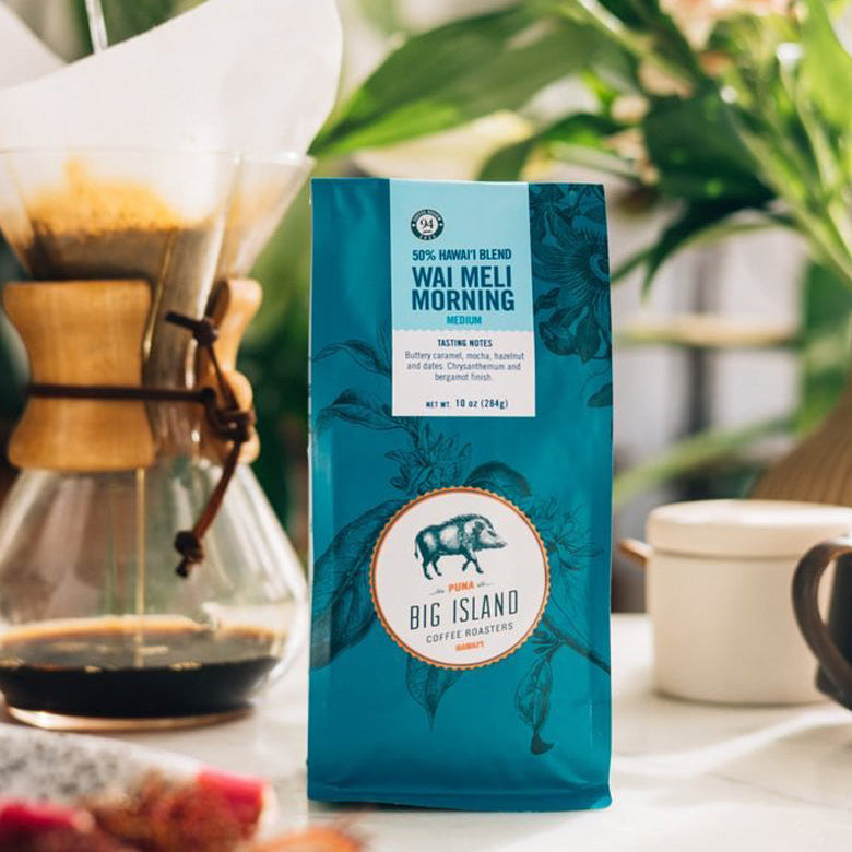 A bag of Big Island Coffee Roasters Wai Meli Morning coffee stands on a table in a bright, real environment with a glass pour-over coffee maker, flowers, and a mug blurred in the background.