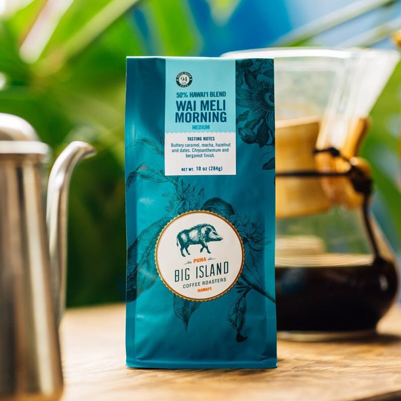 A bag of Big Island Coffee Roasters Hawaiian Blend coffee Wai Meli Morning coffee stands on a wooden table with a coffee pour-over setup and blurred tropical greenery in the background.